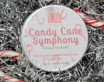 May include: A round tin of Candy Cane Symphony Holiday Tea Blend from Ash Hollow Tea Company. The tin is white with red and white candy cane stripes and features a small illustration of a house. The text on the tin reads "Candy Cane Symphony Holiday Tea Blend" and "Black ceylon tea, peppermint leaves, & candy cane pieces". The net weight is 2 oz by volume.