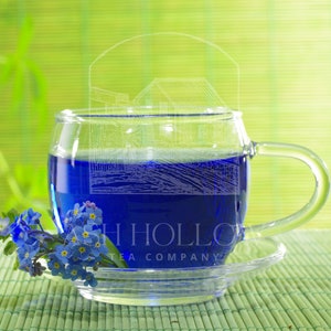 May include: A glass cup filled with blue tea, sitting on a bamboo mat with a small bouquet of blue forget-me-not flowers. The cup has the text "Ash Hollow Tea Company" printed on the side.