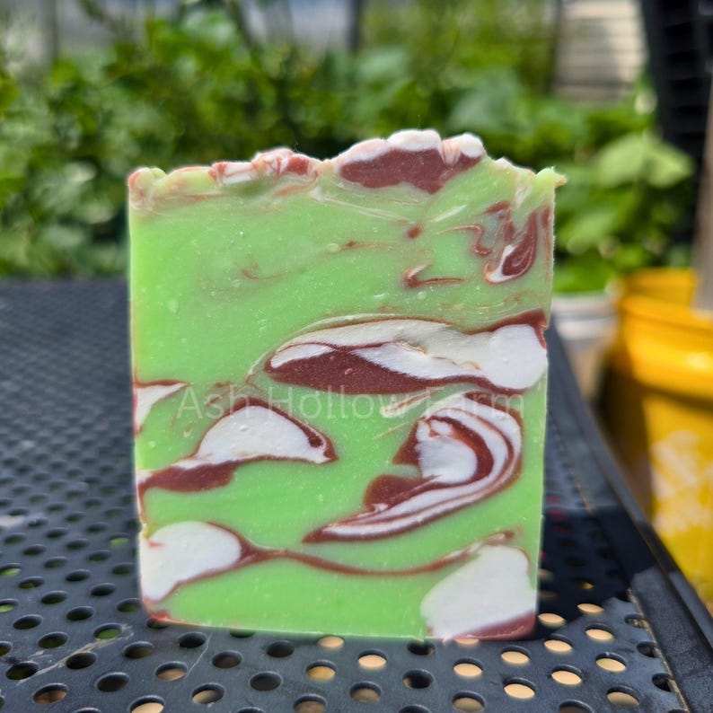 May include: A rectangular bar of handmade soap with a vibrant green base, swirled with white and maroon patterns. The text "Ash Hollow Farm" is visible on the front. The soap has a textured, natural appearance.