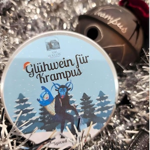 May include: A round, blue and white label with a cartoon image of a Krampus and a small blue creature. The label reads "Glühwein für Krampus" and "Spiced" at the bottom. The label is surrounded by silver tinsel.