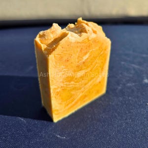 May include: A rectangular bar of handmade soap with a textured, uneven top. The soap is a warm orange color with a marbled pattern and the text "Ash Hollow Farm Soaps" is visible on the side. The soap is on a blue surface.