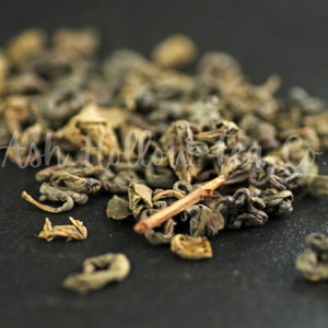 May include: Close-up of loose leaf green tea with a brown twig. The tea leaves are dark green and brown, and some are curled into small balls.