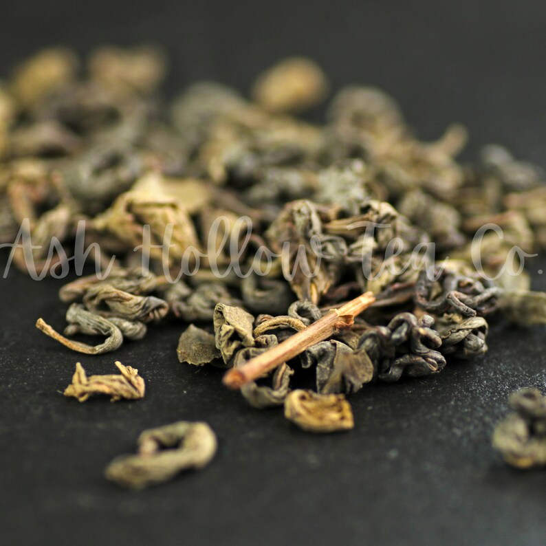 May include: Close-up of loose leaf green tea with a brown twig. The tea leaves are dark green and brown, and some are curled into small balls.