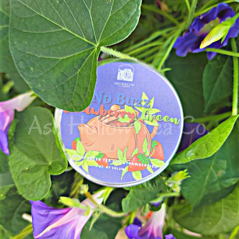 May include: A round, silver tin with a blue label that says "No Buzz Strawberry Green Tea" with a cartoon image of a teapot and strawberries. The tin is surrounded by green leaves and purple flowers.