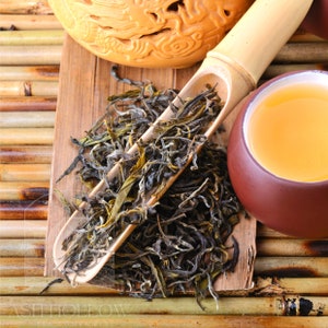 May include: A close-up of loose leaf green tea in a bamboo scoop, with a brown teacup and a ceramic pot in the background. The tea is spread out on a wooden surface with bamboo slats.