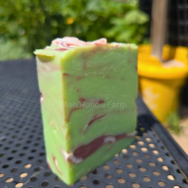 May include: A rectangular bar of handmade soap with a green base color and red and white marbling. The text "Ash Hollow Farm" is visible on the side of the soap. The top of the soap has a pinkish hue.