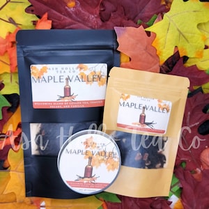 May include: An assortment of tea products from Ash Hollow Tea Co. featuring the "Maple Valley" blend. The image shows a black pouch, a tan pouch, and a round tin, all with labels. The labels display a bottle illustration and the tea blend's ingredients.