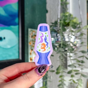 May include: A sticker featuring a cartoon lava lamp design. The lamp is lavender with a blue base and contains three colorful worm-like creatures in orange, purple, and blue. The background is white with green dots.