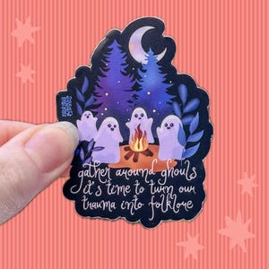May include: A die-cut sticker featuring four white ghosts sitting around a campfire in a dark forest with a crescent moon above. The text reads, "gather around ghouls it's time to turn our trauma into folklore" in white cursive script lettering.