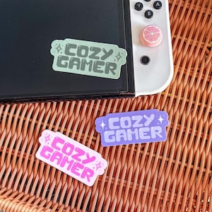 May include: Three stickers with the text "Cozy Gamer" in a pixelated font. The stickers are in different colours: green, purple, and pink. The stickers are on a woven wicker surface.