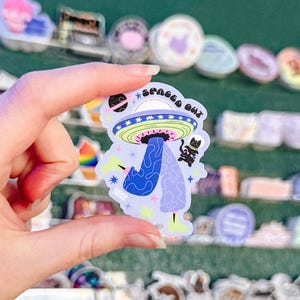 May include: A colorful sticker featuring a mushroom-shaped design with a UFO on top and the text "SPACED OUT". The sticker has a blue and purple color scheme with a black cat holding a star.