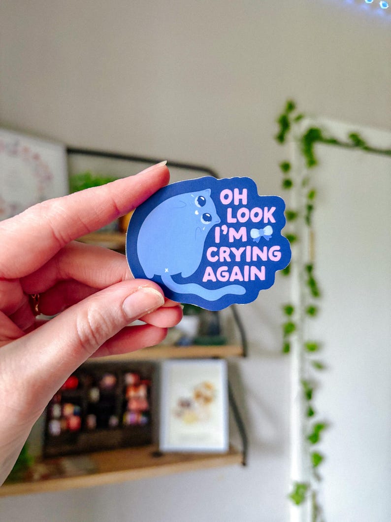 May include: A blue and pink sticker with a cartoon cat and the text "Oh look I'm crying again".