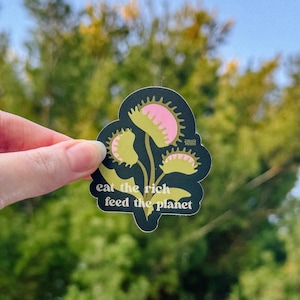 May include: A black sticker featuring a Venus flytrap design with pink and yellow details. The sticker has the text "eat the rich feed the planet" in white. The sticker is held against a blurred green and blue background.