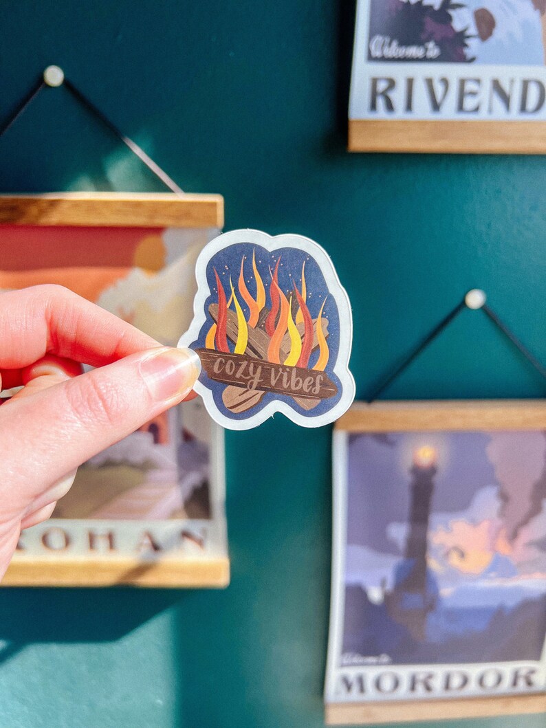 K&ouml;nnte beinhalten: Ein transparenter Aufkleber mit einer Lagerfeuer-Illustration und dem Text "cozy vibes". Die Flammen sind orange und gelb, und die Holzscheite sind braun. Der Aufkleber befindet sich auf einem blauen Hintergrund.