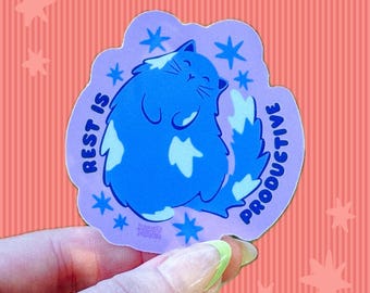 May include: A die-cut sticker featuring a fluffy blue cat with white patches, surrounded by blue star icons on a light purple background. The text REST IS PRODUCTIVE is printed in dark blue capital letters along the curved edges of the sticker.