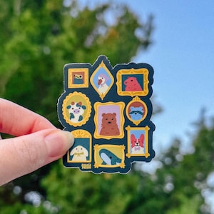 May include: A sticker featuring a collection of framed animal portraits. The sticker includes illustrations of a bear, whale, raccoon, and other animals in ornate gold frames against a dark blue background. The sticker is held by a hand.