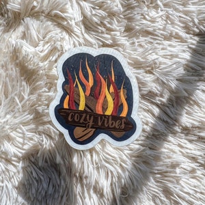K&ouml;nnte beinhalten: Ein Aufkleber mit einer Lagerfeuer-Illustration und dem Text "cozy vibes" in einer handschriftlichen Schriftart. Die Flammen sind orange, gelb und rot, und der Hintergrund ist dunkelblau mit wei&szlig;en Sternen.
