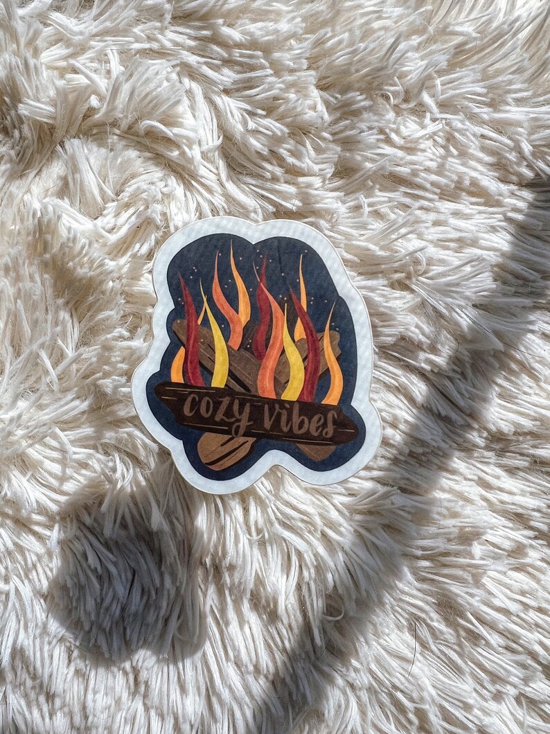 K&ouml;nnte beinhalten: Ein Aufkleber mit einer Lagerfeuer-Illustration und dem Text "cozy vibes" in einer handschriftlichen Schriftart. Die Flammen sind orange, gelb und rot, und der Hintergrund ist dunkelblau mit wei&szlig;en Sternen.