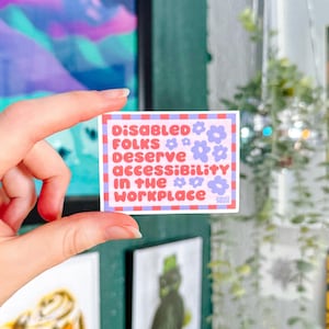 May include: A square sticker with the text "DISABLED FOLKS DESERVE ACCESSIBILITY IN THE WORKPLACE" in red and purple. The sticker has a white border with small flower illustrations. The sticker is held by a hand.