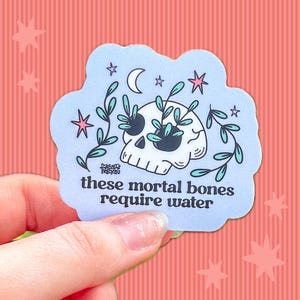 May include: A light blue die-cut sticker featuring a white skull with dark eye sockets, from which green leafy vines emerge. Above the skull are a crescent moon and pink stars. The text reads: these mortal bones require water.