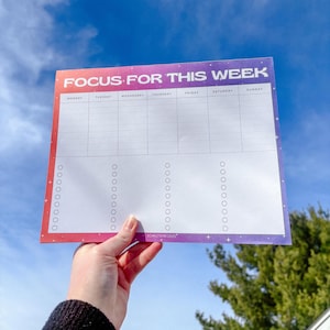 May include: A weekly planner with a gradient of red to purple, held against a blue sky. The planner has the text "FOCUS FOR THIS WEEK" at the top, with days of the week listed below. Check boxes and lines are present for daily tasks.