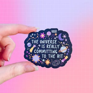 May include: A black sticker with a cosmic design and the text "THE UNIVERSE IS REALLY COMMITTING TO THE BIT." The sticker features stars, planets, and a crescent moon. The sticker is held against a pink and purple gradient background.
