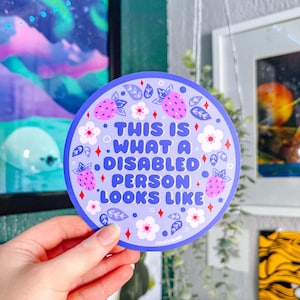 May include: A circular sticker with the text "THIS IS WHAT A DISABLED PERSON LOOKS LIKE" in blue. The sticker has a light purple background with strawberry and floral designs. The sticker is held by a hand.