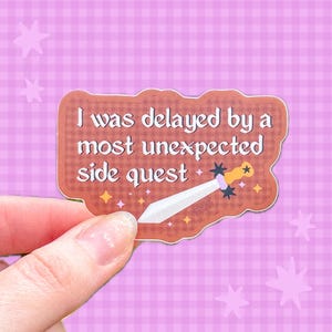 May include: A die-cut sticker with a reddish-brown checkered background featuring the white text I was delayed by a most unexpected side quest and a graphic of a sword with a yellow hilt and a white blade decorated with small yellow stars.
