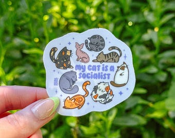 Może przedstawiać: Biała naklejka z wyciętym konturem przedstawiająca różne ilustrowane koty w różnych kolorach i pozach, z fioletowym napisem "my cat is a socialist" pośrodku, otoczona małymi niebieskimi gwiazdkami na jasnoniebieskim tle.