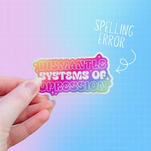 May include: A colorful sticker with the text "Dismantle Systems of Oppression" in a rainbow gradient. The sticker has a white outline and is held by a hand against a blue and pink grid background. The text "Spelling Error" is written above the sticker.