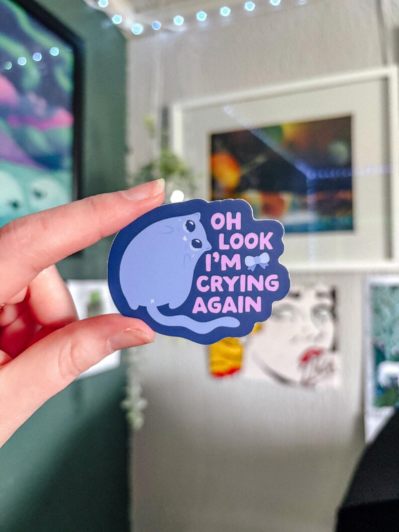 May include: A blue and pink sticker with a cartoon cat and the text "Oh look I'm crying again".