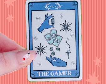 May include: A rectangular sticker featuring a blue and white tarot card design titled THE GAMER. The illustration includes two blue hands, three small cards with celestial symbols, a sword, dice, and star icons on a light background with a blue border.