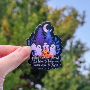 May include: A sticker featuring a nighttime scene with ghosts around a campfire under a crescent moon. The sticker has text that reads "gather around ghouls, it's time to turn our trauma into folklore."