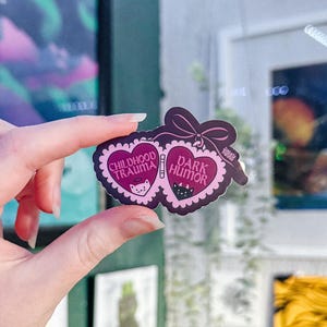 May include: A sticker with a dark pink heart-shaped design featuring the words "CHILDHOOD TRAUMA" and "DARK HUMOR". The sticker has a bow at the top and small cat illustrations. The sticker is held by a person.