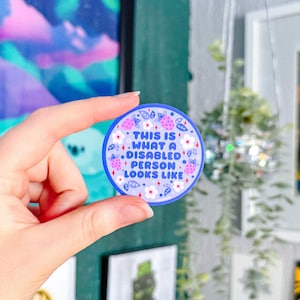 May include: A round sticker with a blue border and the text "THIS IS WHAT A DISABLED PERSON LOOKS LIKE." The sticker has a white background with pink strawberries and white flowers.
