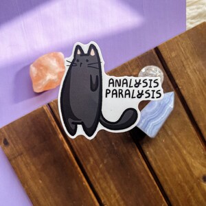 May include: Black cat sticker with the text "Analysis Paralysis" on a wooden surface.