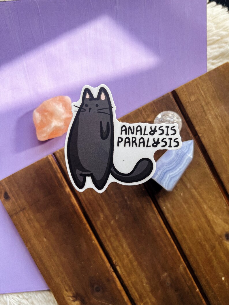 May include: Black cat sticker with the text "Analysis Paralysis" on a wooden surface.