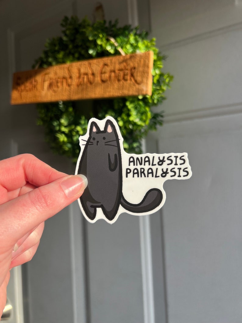 May include: Black cat sticker with the text "ANALYSIS PARALYSIS" in white. The sticker is held in a hand against a gray door.