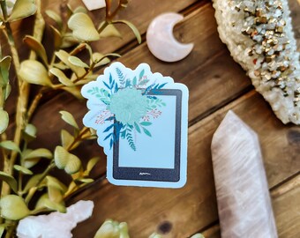 May include: A sticker of a black e-reader with a floral design on the screen. The sticker is on a wooden surface with crystals and greenery.