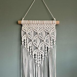Puede incluir: Un tapiz de macramé blanco con un patrón geométrico. El tapiz está sujeto a una varilla de madera y tiene una forma triangular.