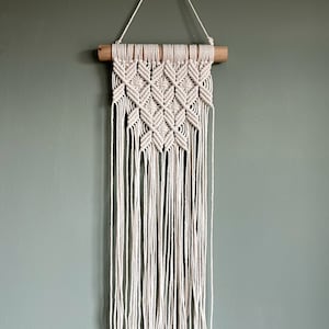 Può includere: Un arazzo da parete in macramè bianco con un motivo geometrico, con un bastone di legno nella parte superiore.