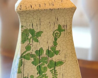 May include: A cream-colored ceramic vase with a crackle finish and green floral designs. The vase has a rectangular shape with a slightly flared top. Black vertical lines add to the design. The floral pattern includes leaves and flowers.
