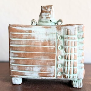 May include: A handcrafted, rectangular ceramic vase with a distressed, aged appearance. The vase features a brown base with horizontal lines and is accented with light blue paint. It has a unique top and small feet.