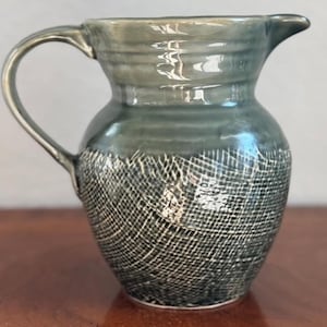 May include: A handmade ceramic pitcher with a dark teal glaze. The upper portion has a smooth finish, while the lower half features a textured, cross-hatched pattern. It has a curved handle and a spout for easy pouring. A decorative piece for the kitchen or dining room.
