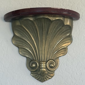 May include: A decorative wall shelf with a dark red, semi-circular wooden top. The shelf's support is a brass-colored, shell-shaped design with intricate scrollwork. The shelf is mounted on a textured white wall.
