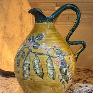 May include: A hand-painted ceramic pitcher with a yellow base and a dark green handle. The pitcher is decorated with a floral and pod design in pink, green, and white. The pitcher has a spout and a curved handle.