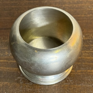 May include: A silver-colored, round napkin ring with a wide opening. The napkin ring has a slightly textured surface and sits on a small, circular base. The object is made of metal and is likely vintage.