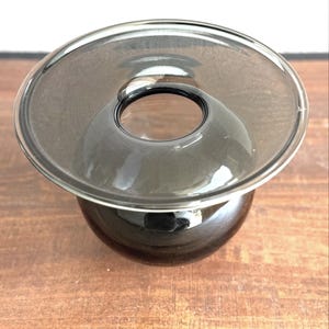 May include: A dark gray glass vase with a wide, flared rim and a rounded base. The vase has a circular opening in the center and sits on a wooden surface. The glass appears to be smooth and reflective, with a slight tint.