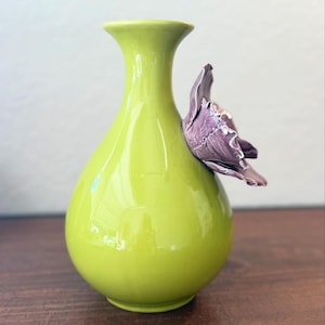 Puede incluir: Un jarr&oacute;n de cer&aacute;mica verde lima con acabado brillante. Una flor morada est&aacute; adherida al lateral. El jarr&oacute;n tiene un cuerpo redondeado y un cuello acampanado. Decoraci&oacute;n del hogar.