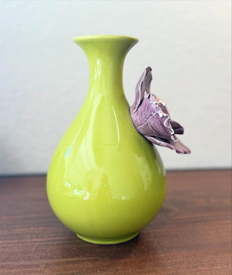 Puede incluir: Un jarr&oacute;n de cer&aacute;mica verde lima con acabado brillante. Una flor morada est&aacute; adherida al lateral. El jarr&oacute;n tiene un cuerpo redondeado y un cuello acampanado. Decoraci&oacute;n del hogar.